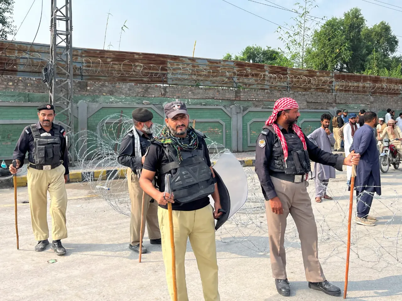 Uniformed police in body armour with batons and shield behind razor wire, bystanders in background.