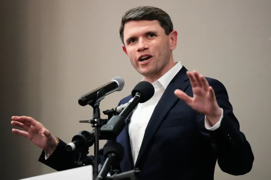 A man in a navy suit speaks at a podium with microphones, gesturing with one hand.