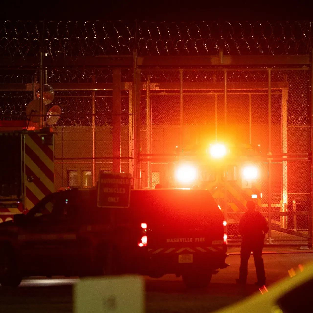 Night view of fire trucks and emergency lights behind a razor-wire prison fence gate.