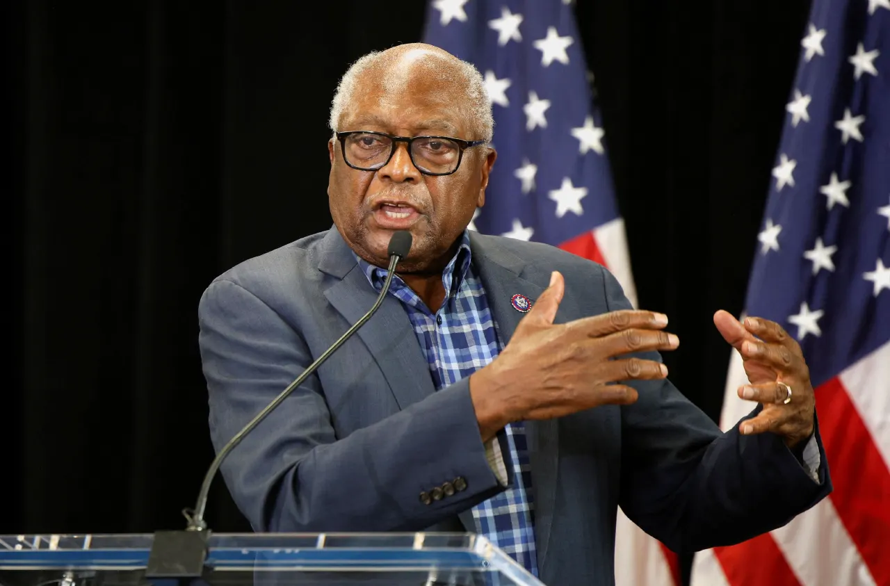 Older man in glasses and blue jacket speaking at a podium before American flags.