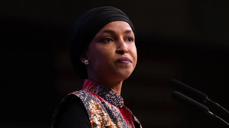 Woman in head covering and patterned top speaking at microphones against dark background