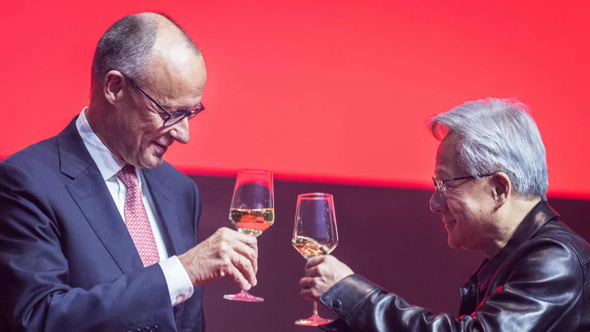 Two men in suits clink wine glasses against a bright red stage backdrop, smiling.