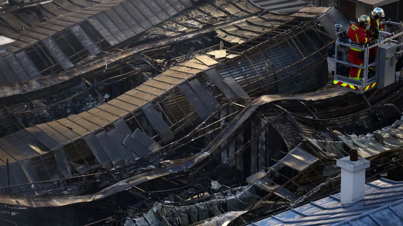 Charred, collapsed market roof with firefighters on an aerial platform inspecting the damage.