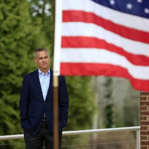 Man in navy blazer stands with hands in pockets beside a blurred American flag.