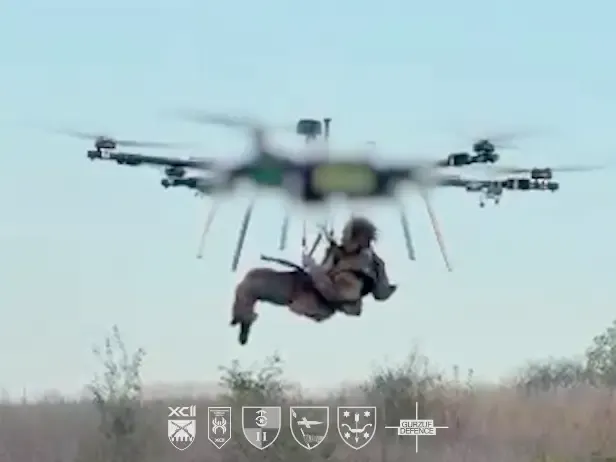 Large multirotor drone lifts a soldier in harness above a grassy field; unit insignia shown below.