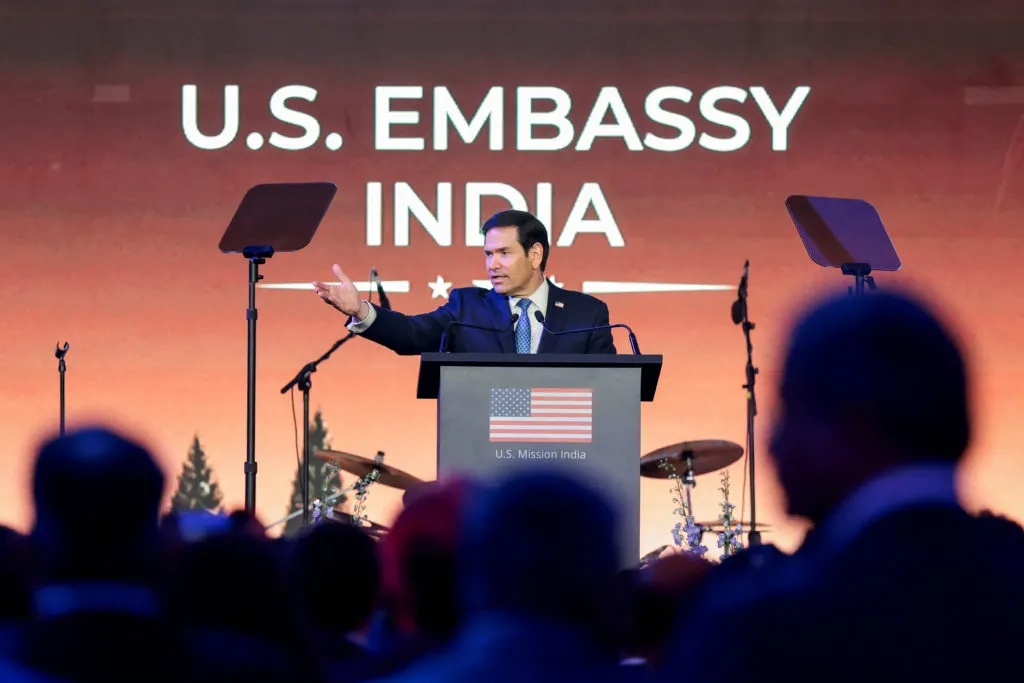 Man in dark suit gesturing while speaking at podium labeled U.S. Embassy India, audience silhouetted.