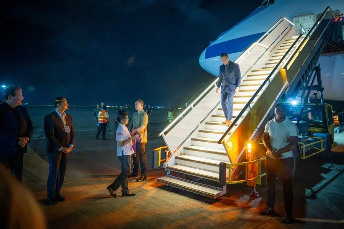 A man in a suit descends an aircraft boarding staircase at night while officials wait on the tarmac.