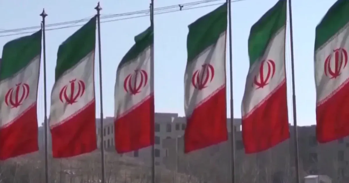 Row of Iranian national flags flying on poles before a building on a hillside.