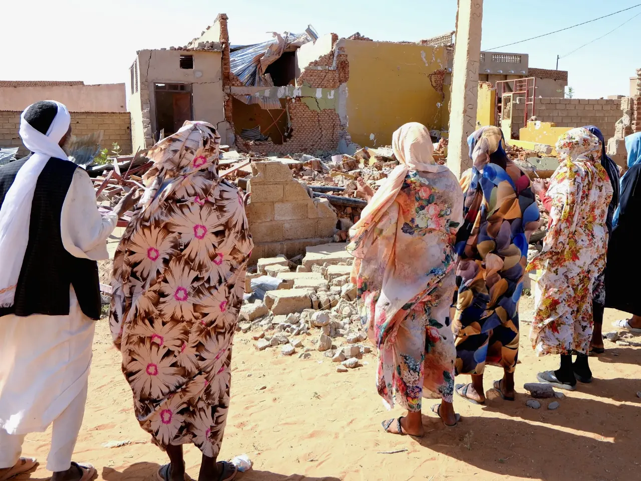 Women and a man in traditional dress view a bombed, rubble-strewn building in Sudan.