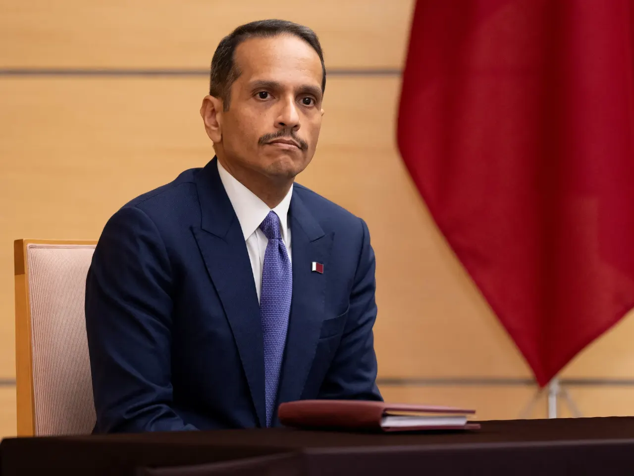 Man in navy suit seated at a table beside a large red flag, with a small flag pin.
