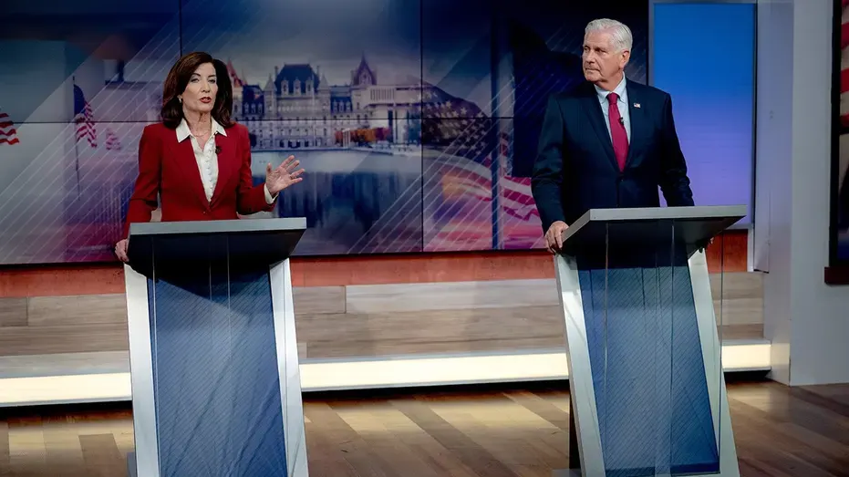 Two candidates stand at lecterns on a studio debate stage, one gesturing mid-speech.