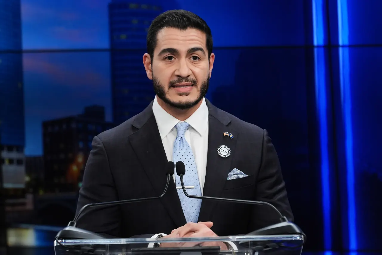 A man in a dark suit and blue tie speaks at a clear podium with microphones before a blue backdrop.