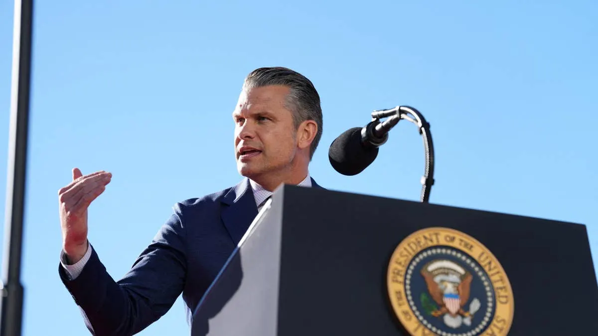 A man in a dark suit speaks at a podium bearing a Presidential seal under blue sky.