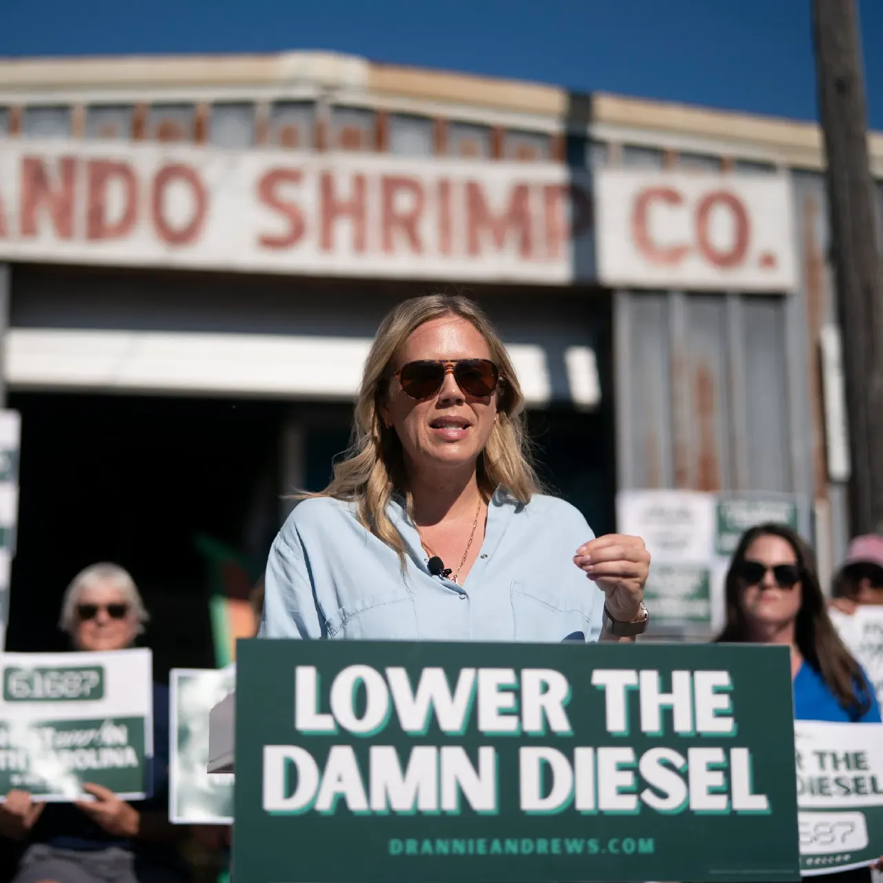 A woman speaks at a podium with a green sign reading Lower the Damn Diesel, supporters behind her.