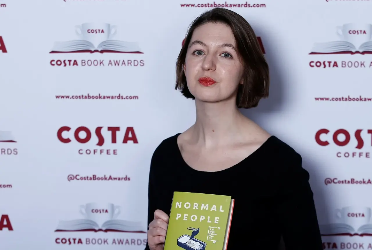 Woman in black top holding a copy of Normal People at Costa Book Awards backdrop.