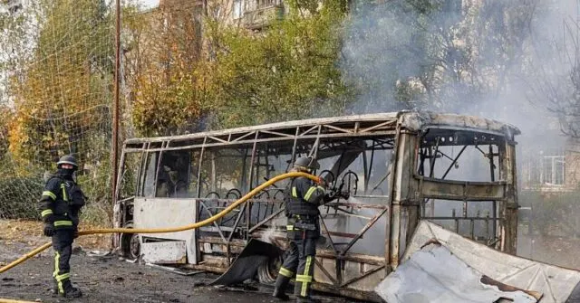 Firefighters hose down a burned-out bus with smoke rising nearby