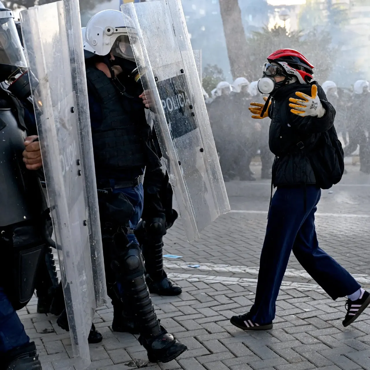 Riot police with shields and helmets face a masked protester in a helmet and gas mask on a city street.