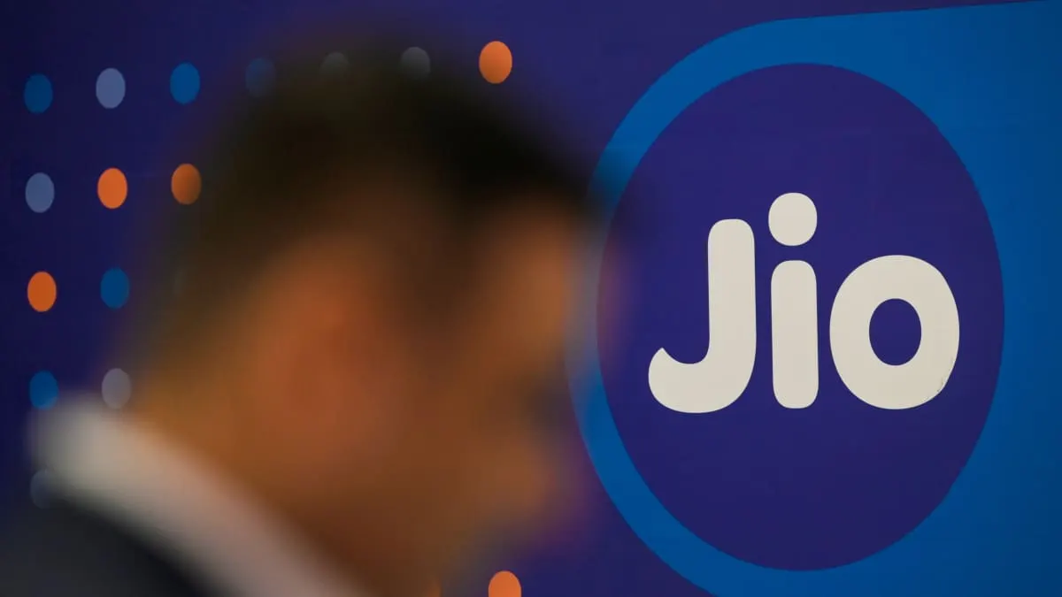 Blurred figure in foreground in front of large illuminated Jio logo on blue backdrop.