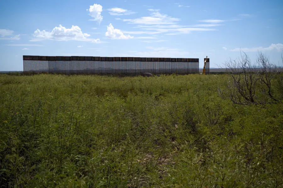 Rusted steel border barrier rising above green scrubland under a partly cloudy blue sky.