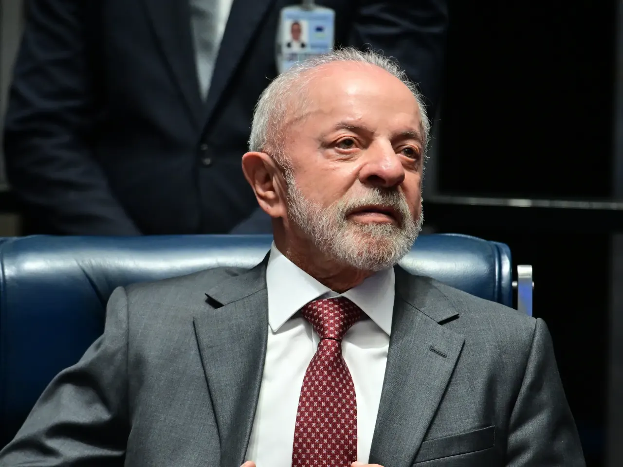 Older bearded man in grey suit and red tie seated in a blue chair, looking to the side.