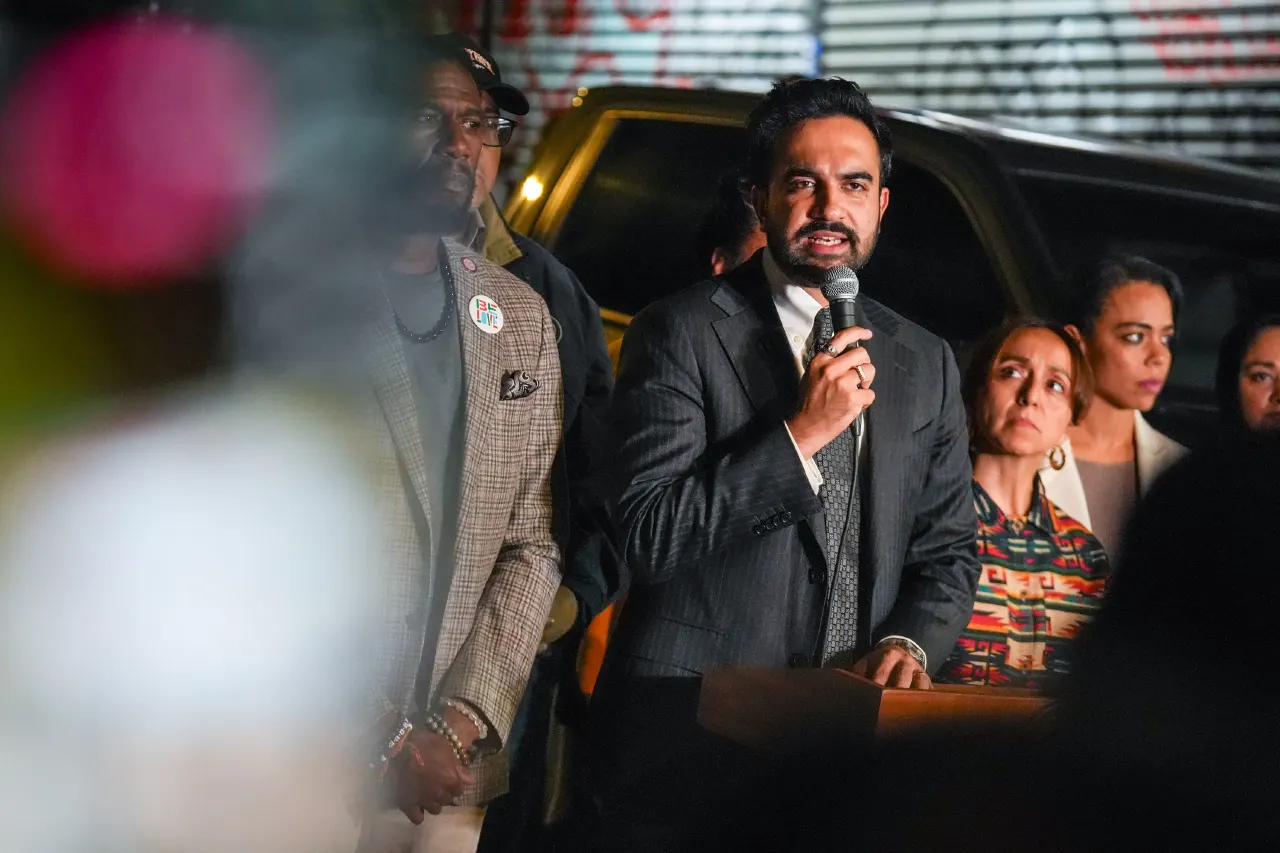 Man in dark suit speaking into microphone at podium, with people and a vehicle behind him at night.