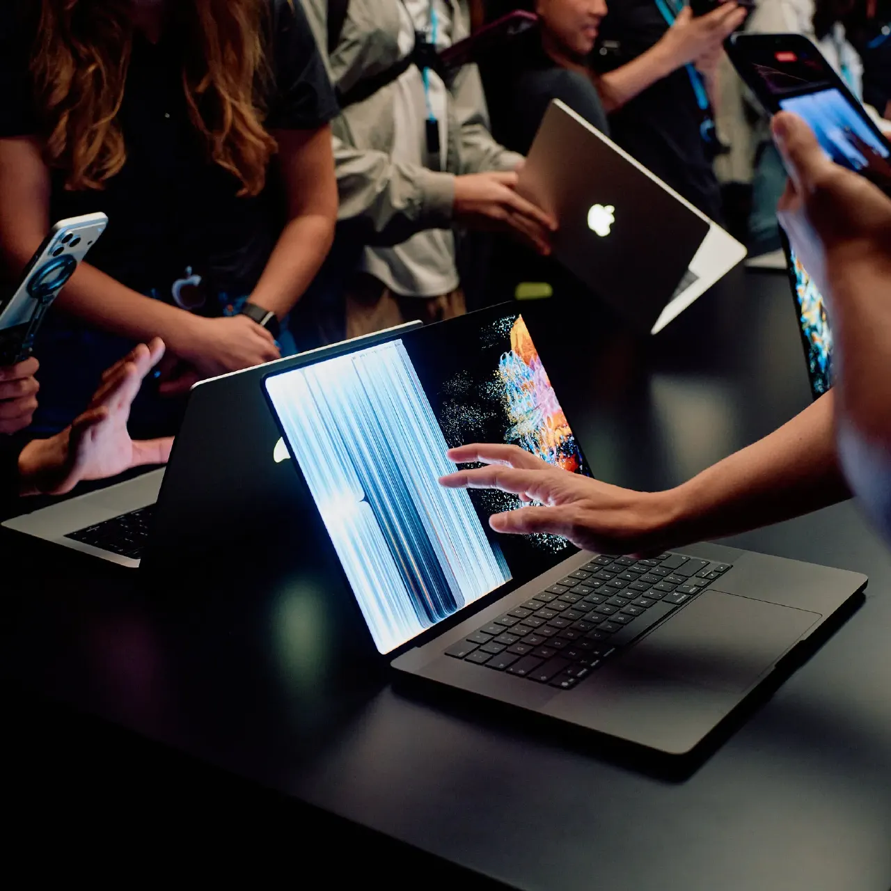 Hands touching a dark Apple laptop screen at a crowded product demonstration event.