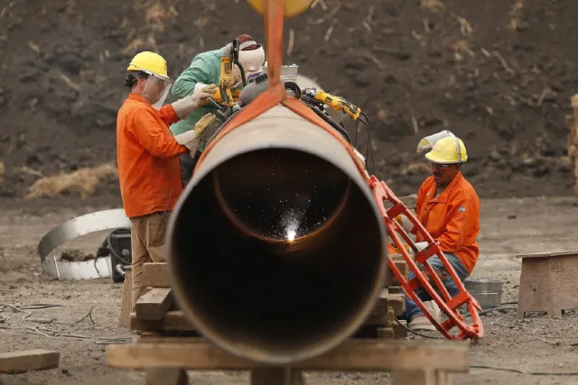 Workers in hard hats welding a large steel pipe segment at a construction site.