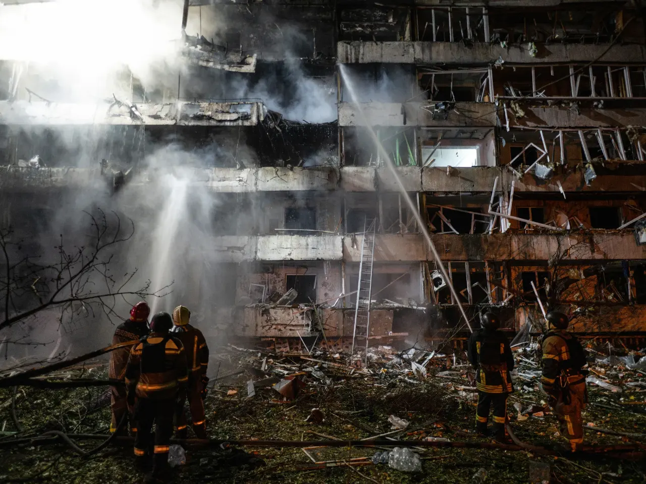 Firefighters spray water at a heavily damaged, smoke-filled residential building amid debris.