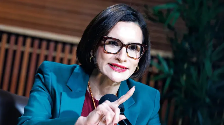 Woman with glasses and dark bob speaking, gesturing, wearing teal blazer, near a microphone.