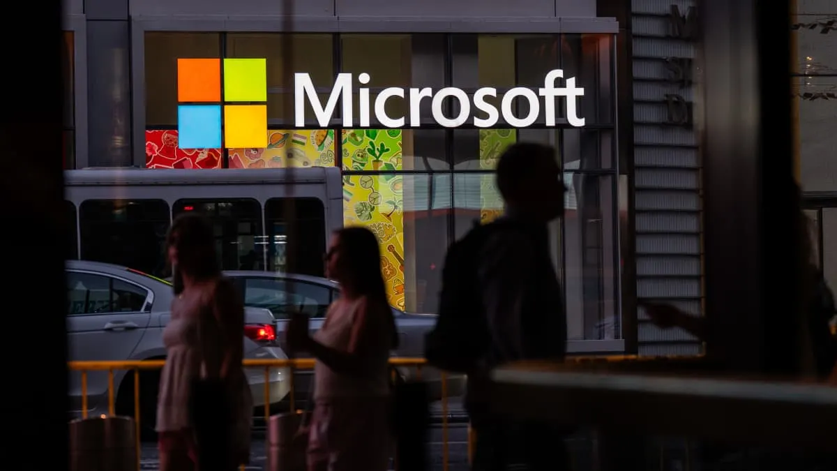 Illuminated Microsoft logo on a building facade, with silhouetted pedestrians passing in front.