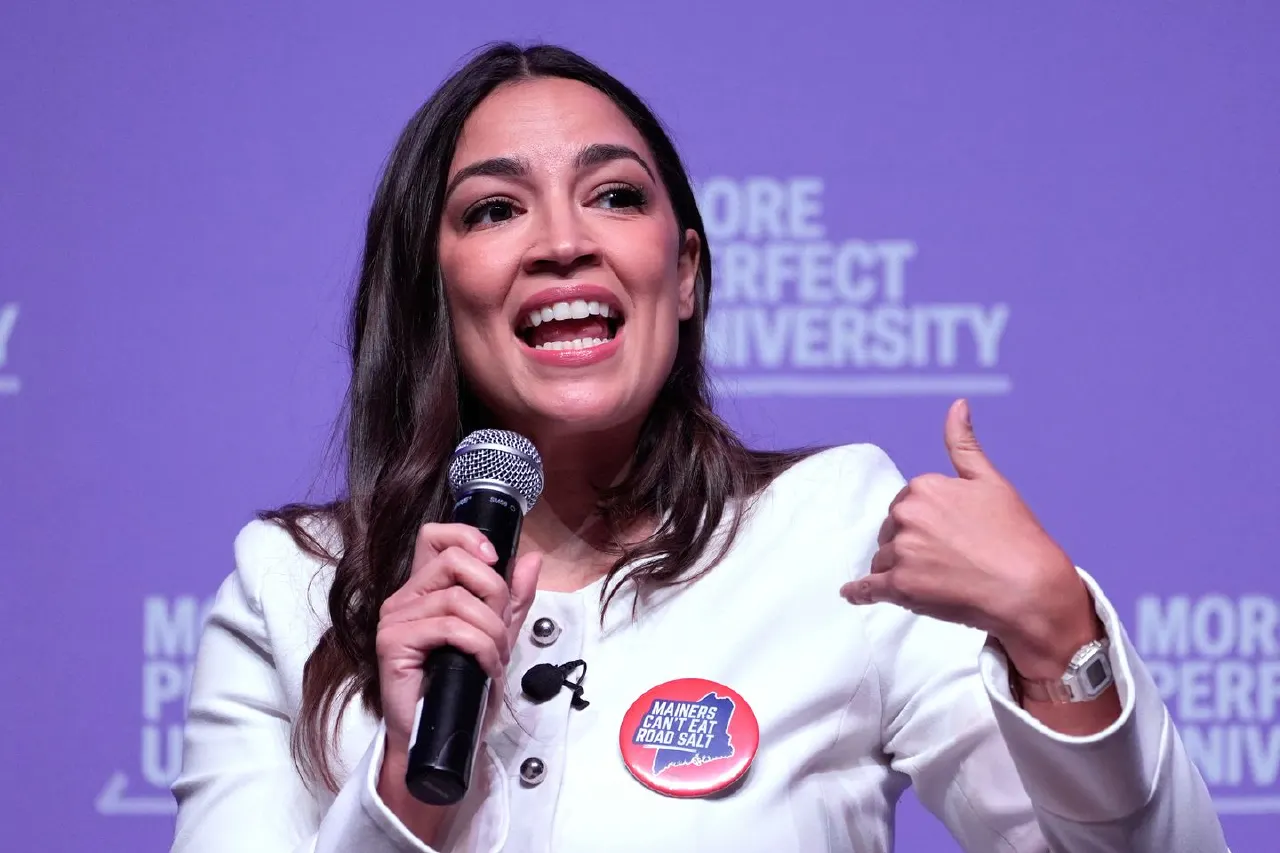 Woman in white blazer speaking into microphone, gesturing, wearing button reading 'Mainers can't eat road salt'.