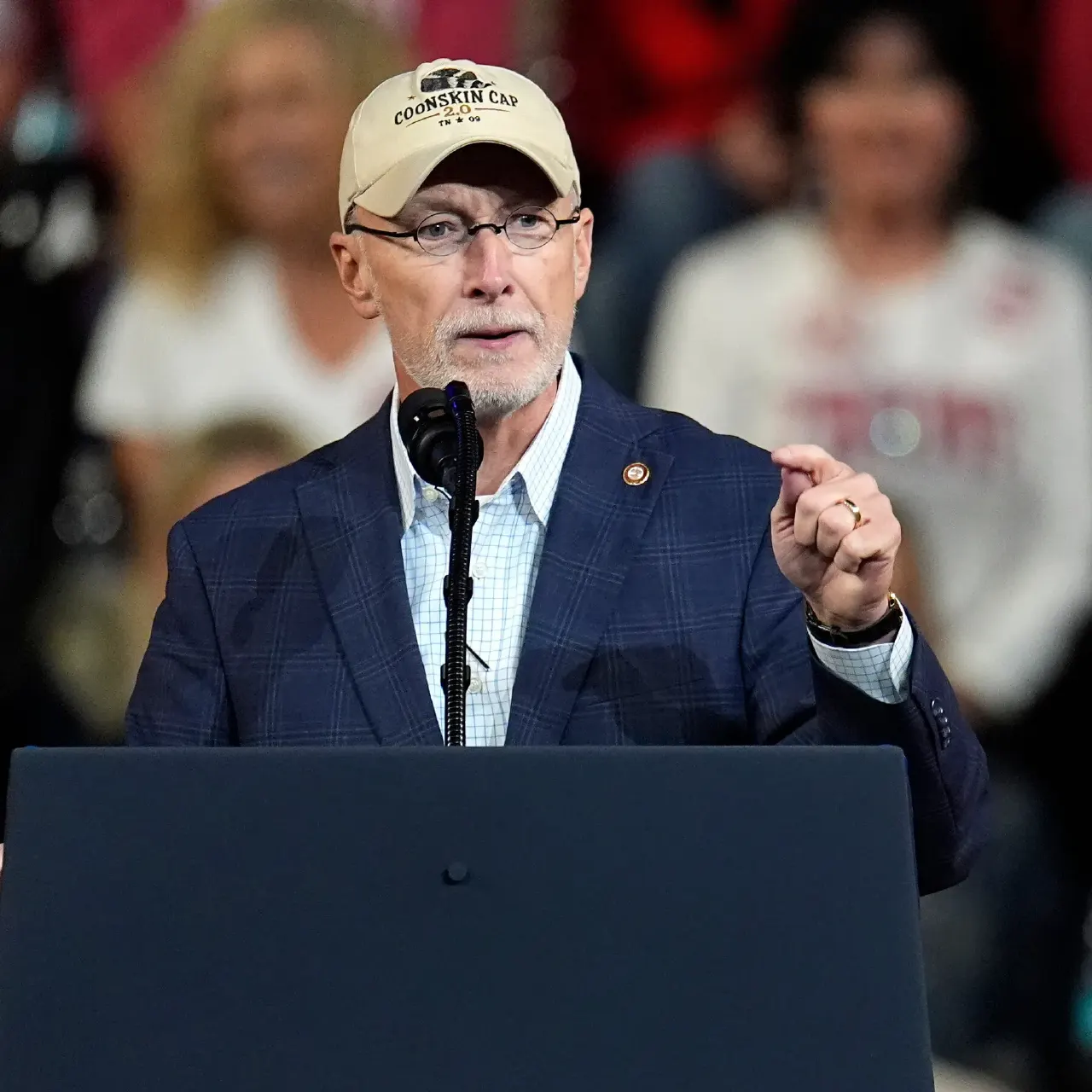 Man in cap and blazer speaking at a podium with a microphone before a crowd.