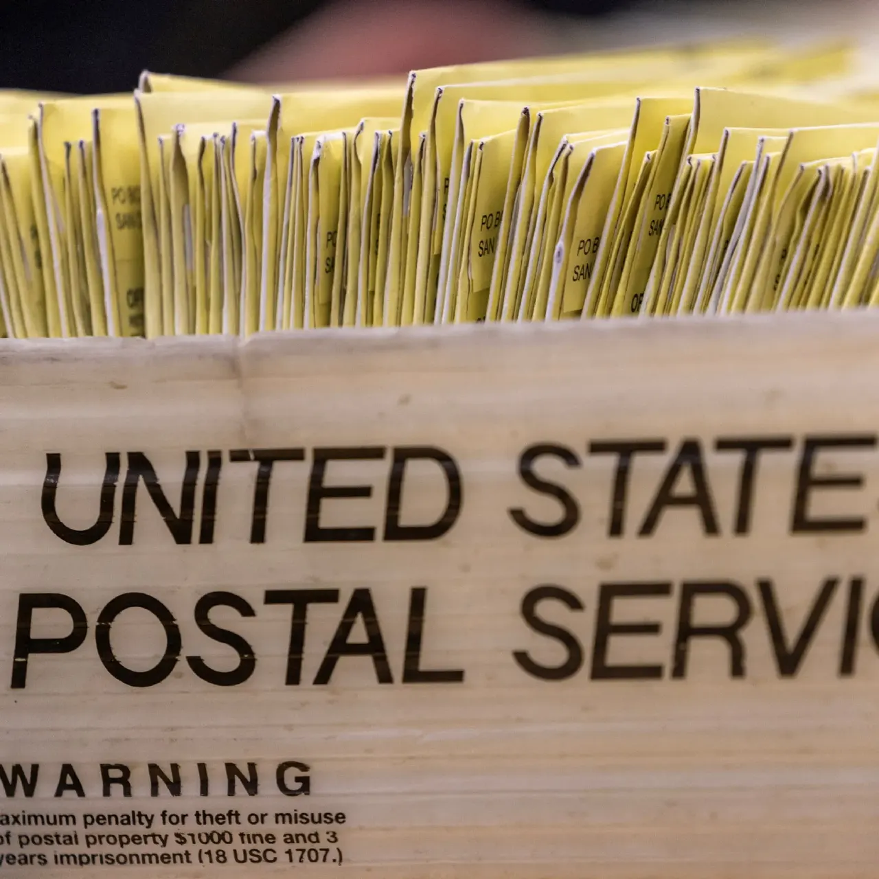 Stacked yellow mail-in ballot envelopes in a United States Postal Service tray, close-up.