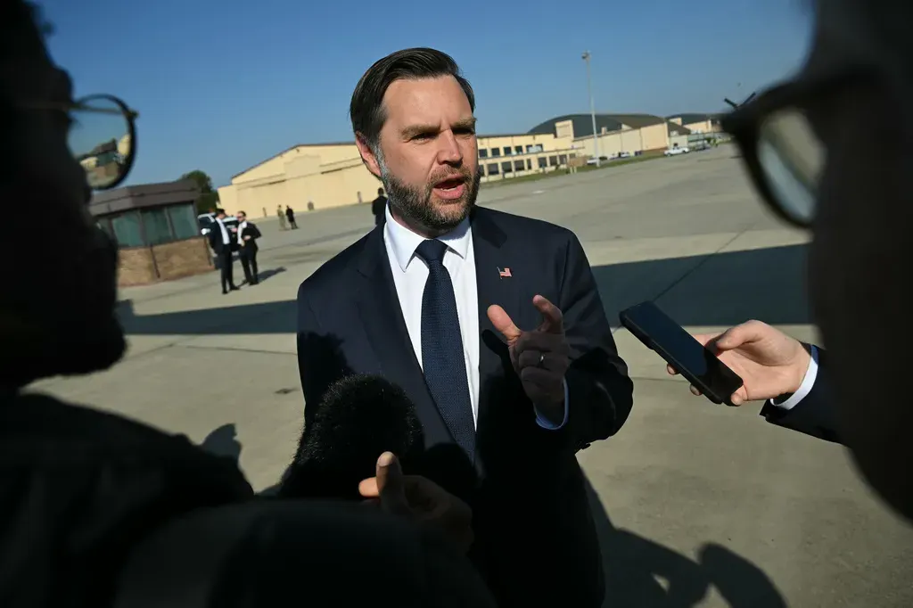 A man in a dark suit speaks to reporters on an airport tarmac, gesturing with his hand.