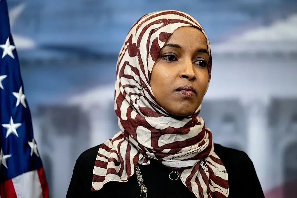 Woman in patterned headscarf and black top beside an American flag, blurred backdrop.