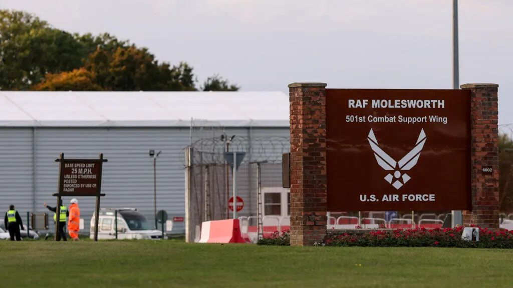 Entrance sign for RAF Molesworth with U.S. Air Force logo, barbed-wire fence, and security personnel.
