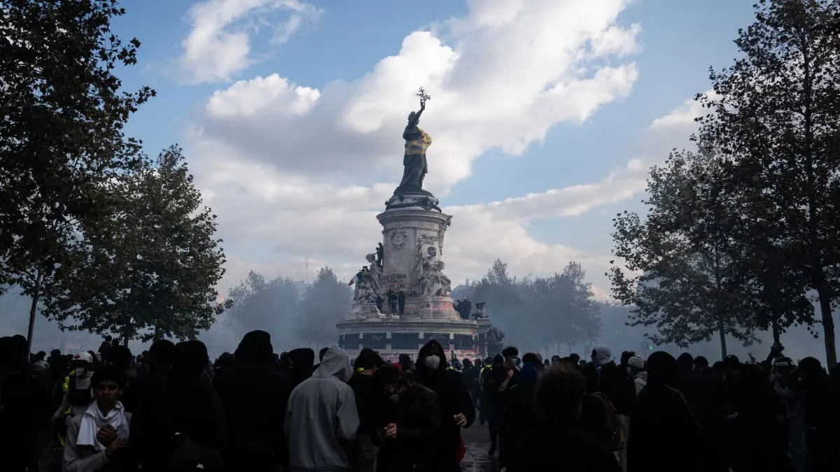 Crowd of masked, hooded demonstrators gathered around a city statue amid smoke and trees.