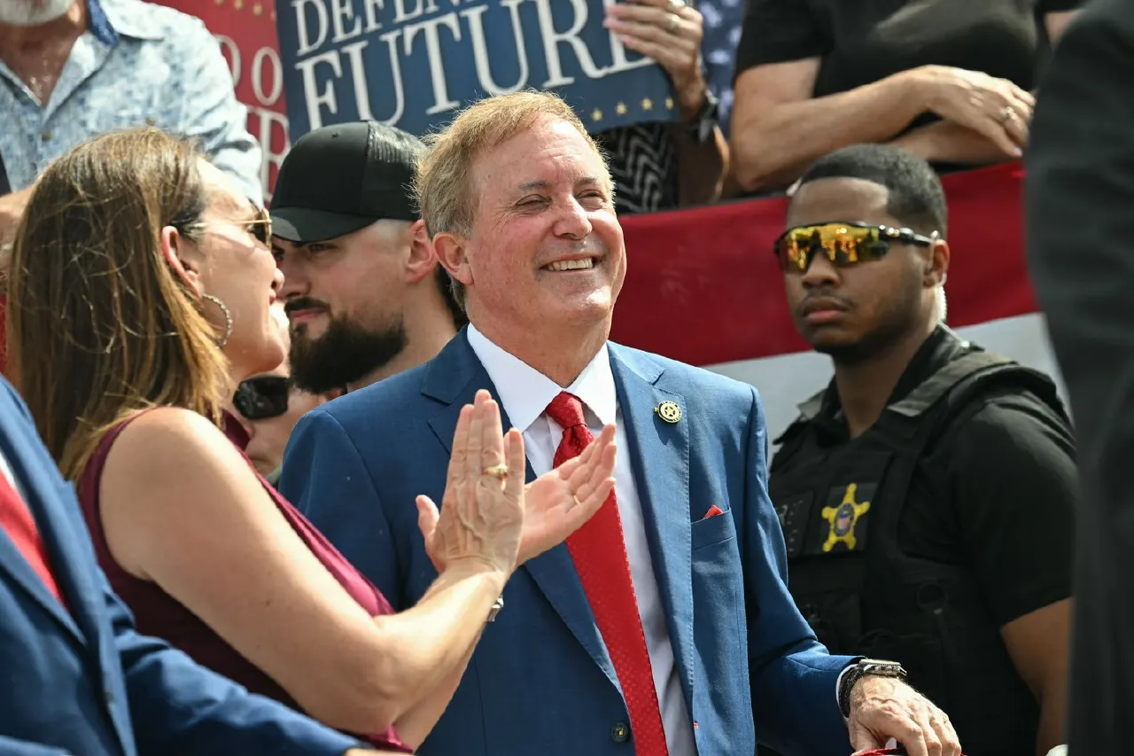 Smiling man in blue suit and red tie applauds at rally; partial campaign sign and flag behind.