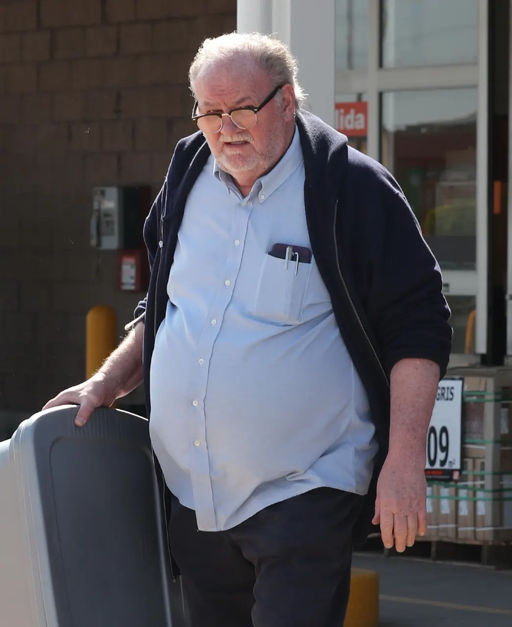 Older man in glasses and light blue shirt walking outside a store, pushing a cart.