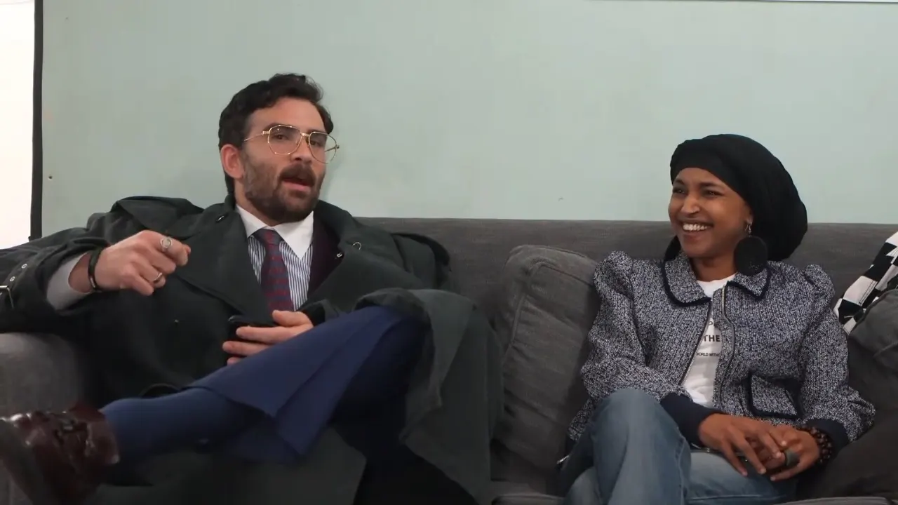 Two people seated on a grey couch in casual conversation, one laughing, one gesturing