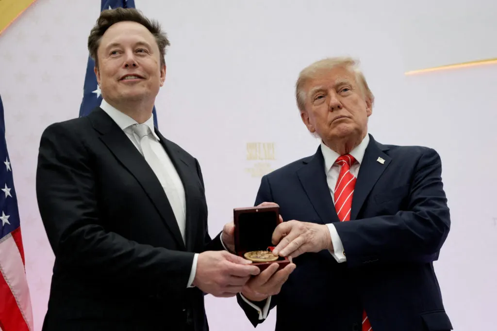 Two men in dark suits exchange a small gold medal in a velvet box, with an American flag behind.