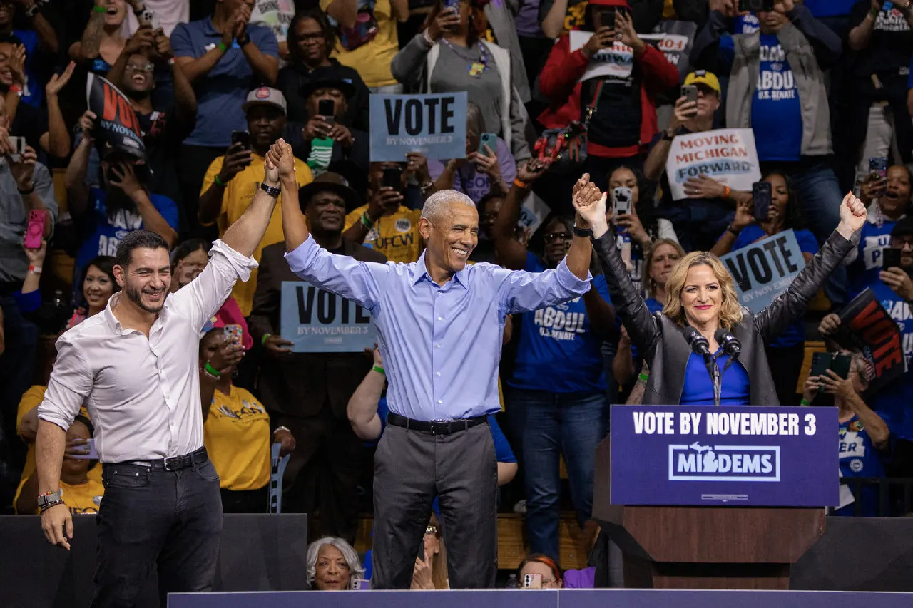 Three smiling people raise joined hands on stage at a rally; crowd holds VOTE signs, podium reads Vote by November 3.