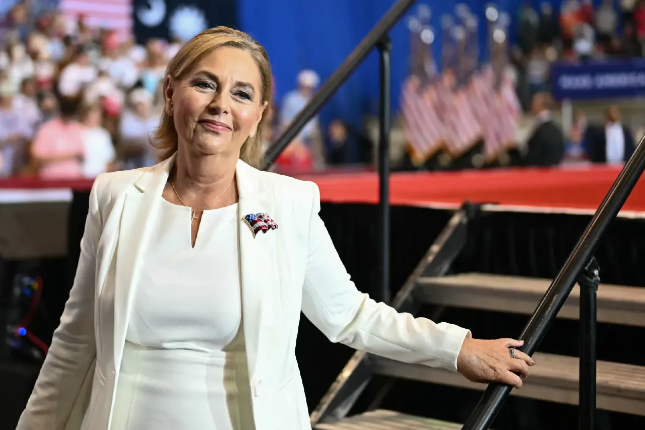Woman in white suit with small flag pin smiles beside stairs at a rally stage.