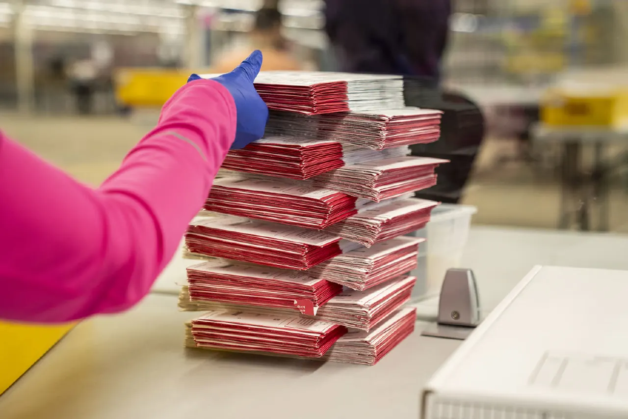 Gloved hand stacking red-envelope ballots on a table in a processing facility