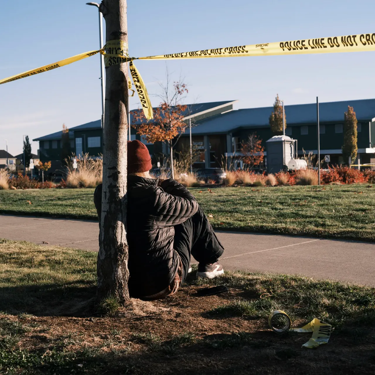 Person sits against a tree behind police tape on a school-area lawn, face hidden.