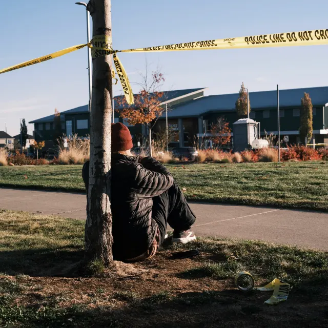 Person sits against a tree behind police tape on a school-area lawn, face hidden.