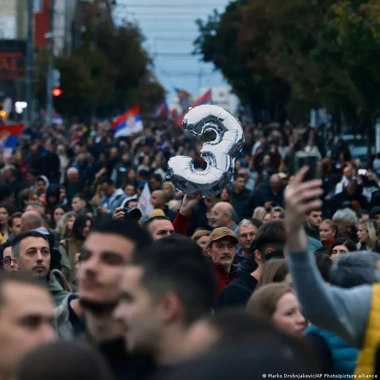 Large crowd marching down a city street, with Serbian flags and a silver balloon numeral held aloft.