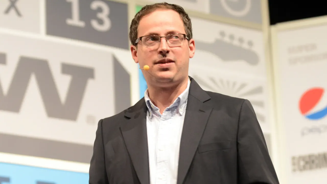 Man in glasses and dark blazer speaking on stage at a conference, with a lapel microphone.