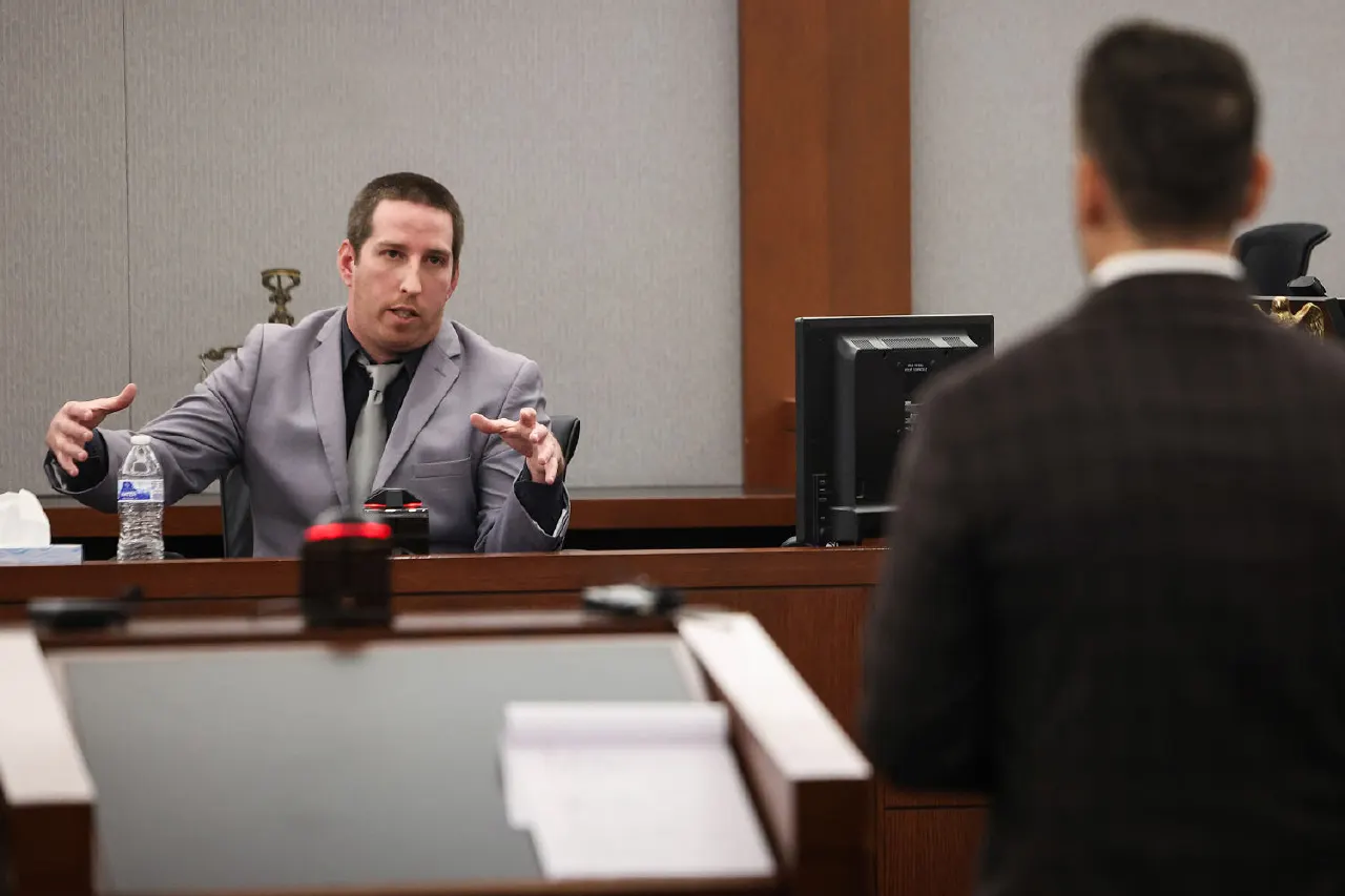 Man in grey suit gestures while speaking in a courtroom, another man with back turned