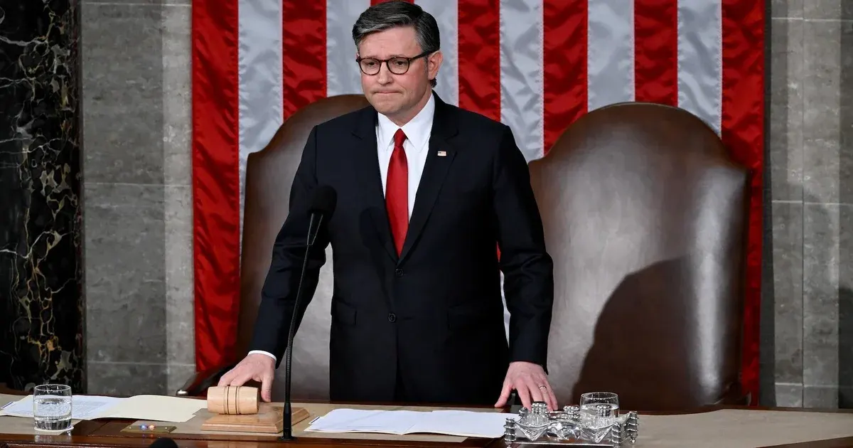 Man in dark suit and red tie stands at a House chamber rostrum beside a gavel and flag.